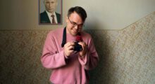 A man in a pink sweater smiles at his camera. A picture of Vladimir Putin is on the wall behind him.