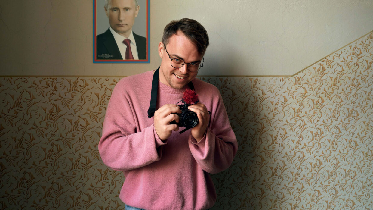 A man in a pink sweater smiles at his camera. A picture of Vladimir Putin is on the wall behind him.