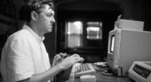 A white man is sitting at an old, CRT-monitor computer.