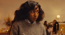 A young Black woman wears a gray sweater and looks off into the corner of a room.