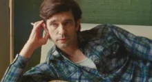 A white man in his 40s with dark hair and a blue plaid shirt leaning on a couch with cigarette