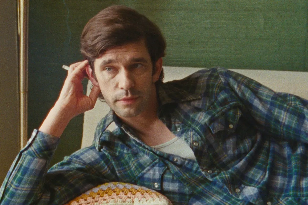 A white man in his 40s with dark hair and a blue plaid shirt leaning on a couch with cigarette