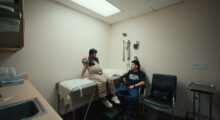 A pregnant woman is sitting on a table in a doctor's office while a man sits next to her on a chair.