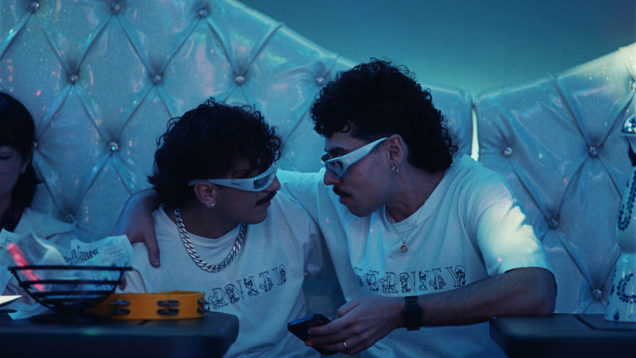 Two men, dressed identically and wearing white paper glasses, look at one another.
