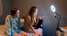 Two young women sit on a bed and look into a phone camera illuminated by a ring light.