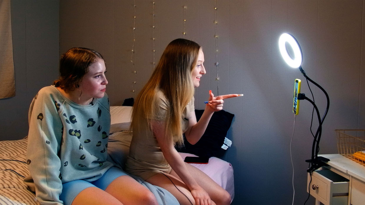 Two young women sit on a bed and look into a phone camera illuminated by a ring light.