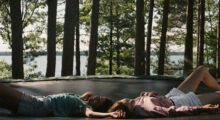 Two young people, one man and one woman, are lying on a trampoline. The ocean is visible in the background behind some trees.