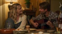 Carey Mulligan and Tom Basden, who is holding a guitar as they sit at a table.