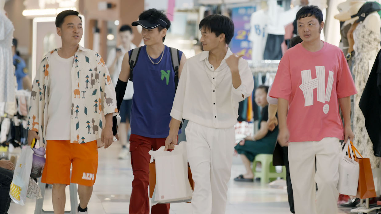 Four young men, all sporting trendy garb, walk through a mall with shopping bags.