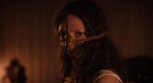 A young woman with long curly hair wearing a facemask looks at the camera.