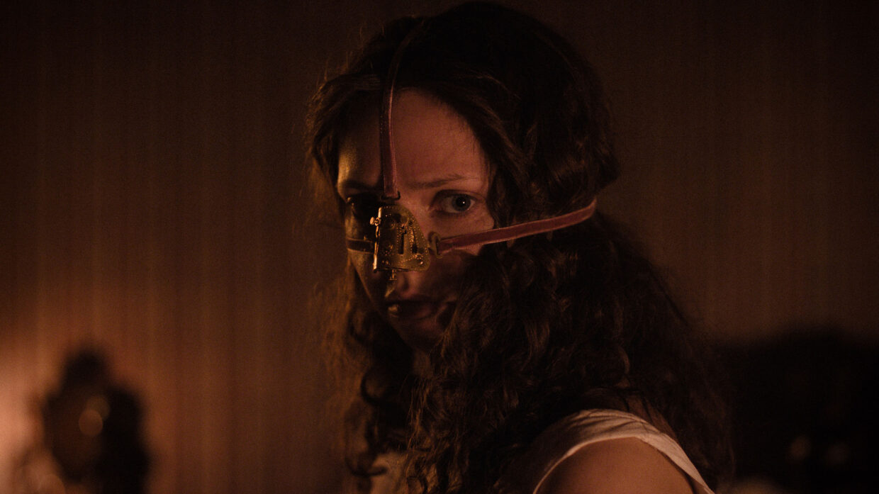 A young woman with long curly hair wearing a facemask looks at the camera.