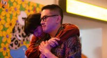 Two men embrace, one wears glasses and the other hugs from behind.
