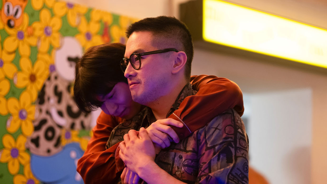 Two men embrace, one wears glasses and the other hugs from behind.