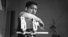 A black and white photo of filmmaker and artist Robert A. Nakamura, who smokes a cigarette while leaning on a camera.