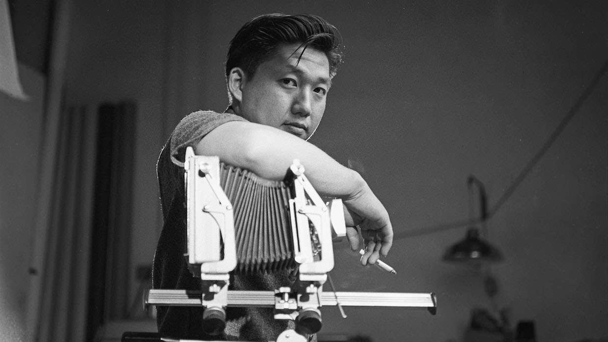 A black and white photo of filmmaker and artist Robert A. Nakamura, who smokes a cigarette while leaning on a camera.