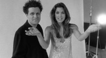 A black and white image of a man with unruly short black hair wearing all black posing next to a long-haired model in a sequin dress.