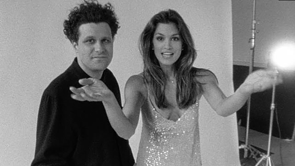 A black and white image of a man with unruly short black hair wearing all black posing next to a long-haired model in a sequin dress.