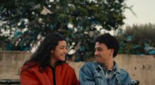 A girl with long, black hair wears a red jacket. She is sitting next to a boy with short curly hair and a blue denim jacket. They look at each other lovingly.