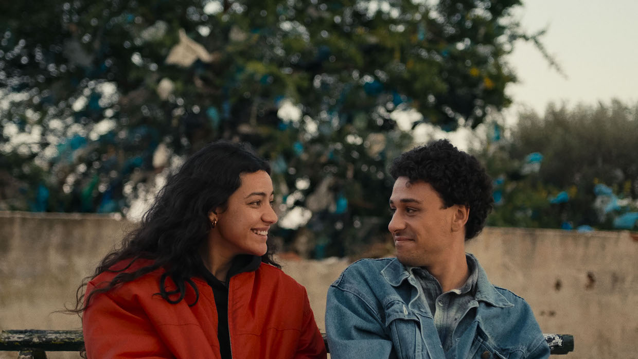 A girl with long, black hair wears a red jacket. She is sitting next to a boy with short curly hair and a blue denim jacket. They look at each other lovingly.