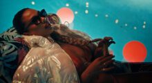 A woman wearing a white puffer jacket, red bikini top and sunglasses reclines amid a neon blue background.