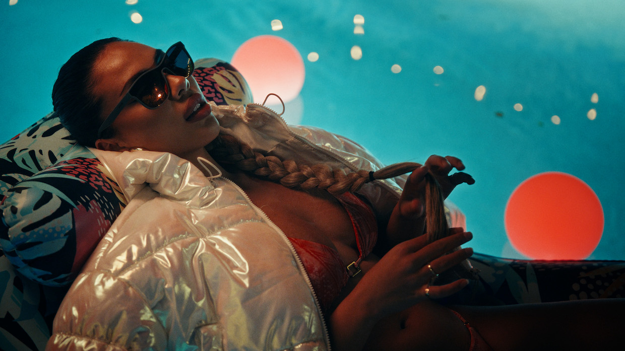 A woman wearing a white puffer jacket, red bikini top and sunglasses reclines amid a neon blue background.