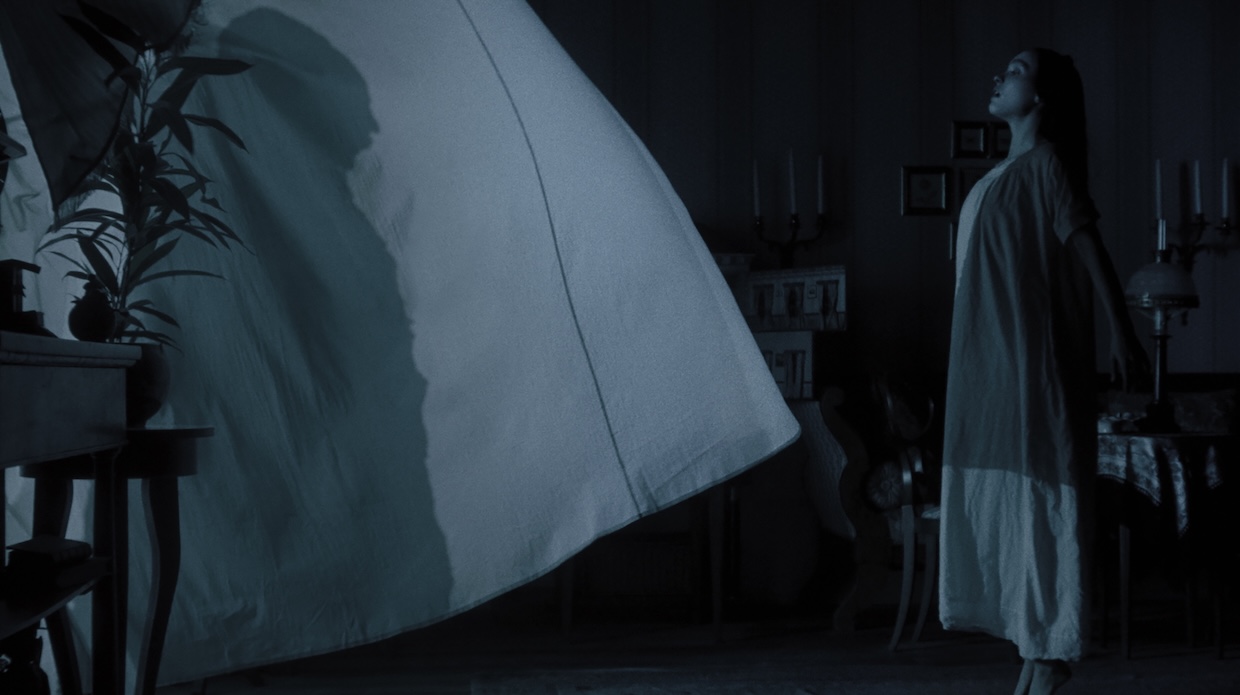 A man's caped silhouette is seen through a white window curtain with a woman standing next to it.