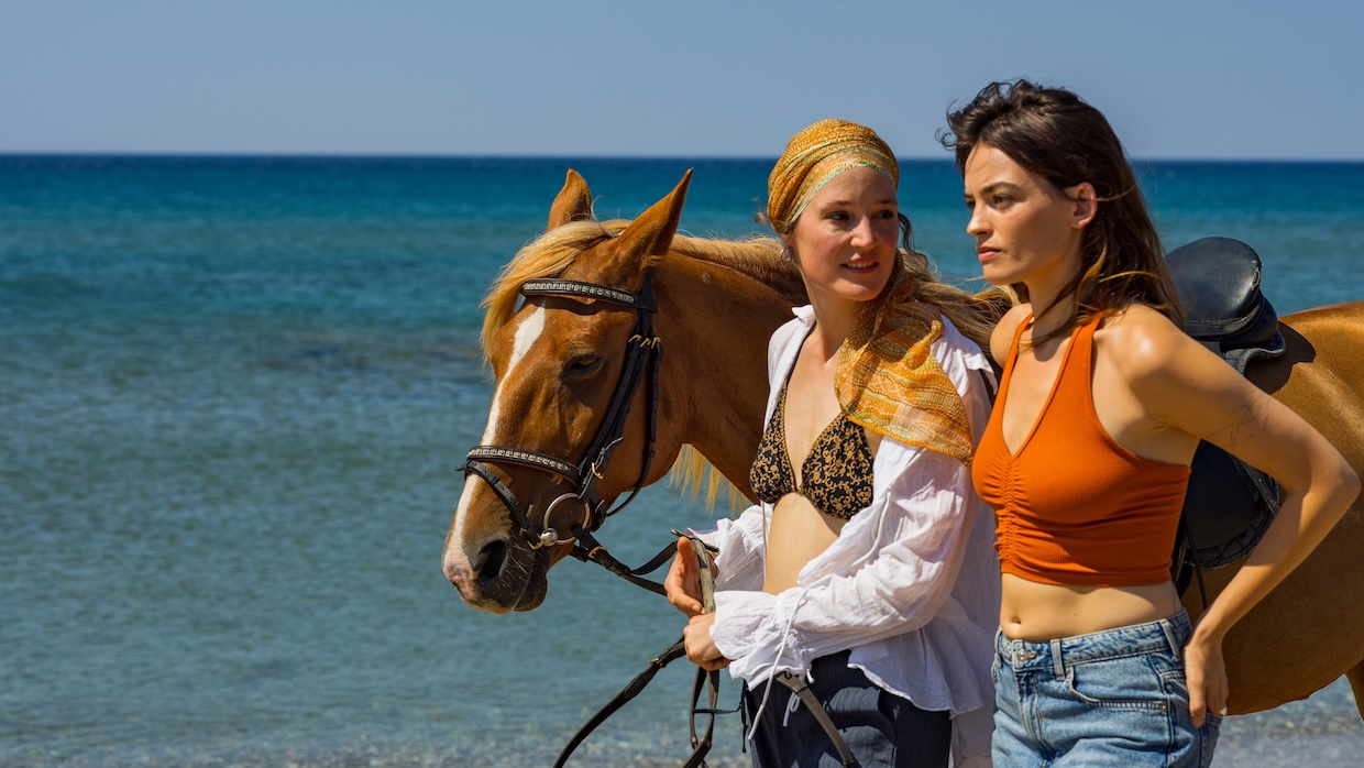 Two women and a horse walk on a sunny beach by bright blue water.