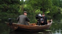An actor in costume sits in a boat opposite a cinematographer with a camera.