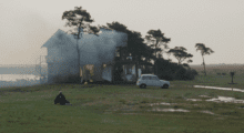 A person sits on a green field in front of a house on fire.