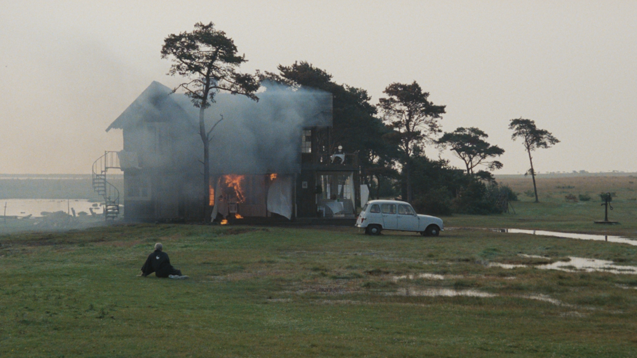 A person sits on a green field in front of a house on fire.