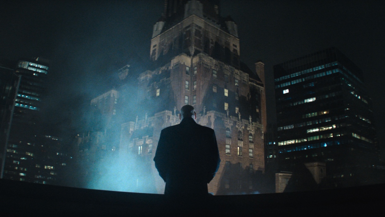 A man stands on a roof in the big city and stares into the night as steam rises before him.