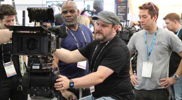 A group of man crowd around a camera on a trade show floor.