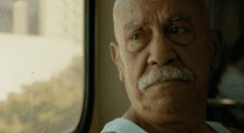 An older Latino man, who is bald and has a white mustache, looks out of a bus window.