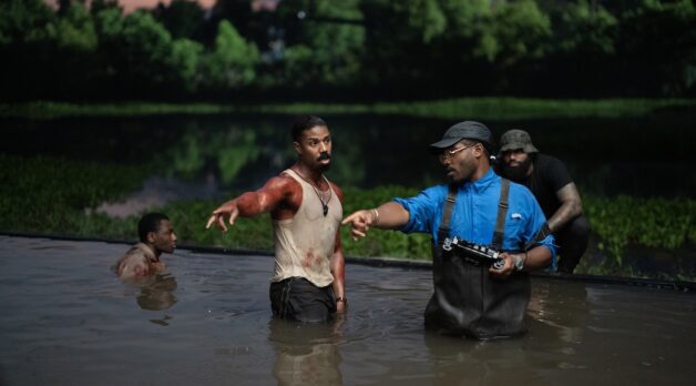 A man in a blood-stained undershirt and a man in overalls stand in a pool of water outdoors.