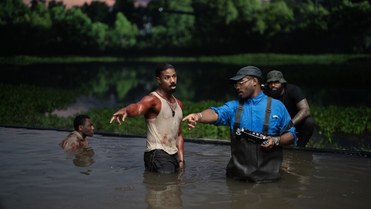 A man in a blood-stained undershirt and a man in overalls stand in a pool of water outdoors.
