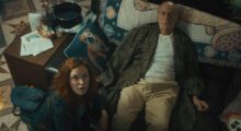 An elderly man lays on a plush couch surrounded by pillows. Kneeling next to him is a younger red-haired woman. They both gaze upward.