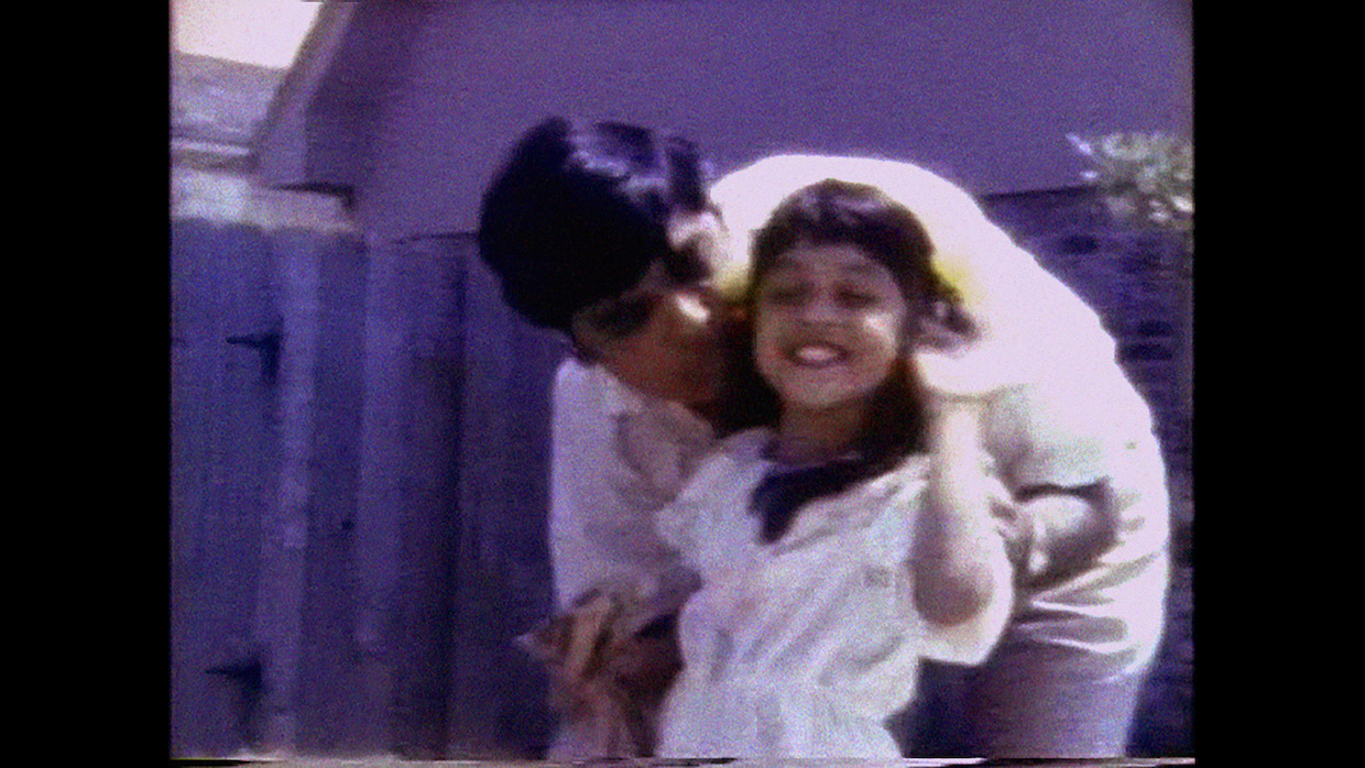 A still from a VHS home movie of an Indian father, with cropped black hair and a white shirt, bending down to kiss his young daughter on the head; she has long black hair and also wears white.
