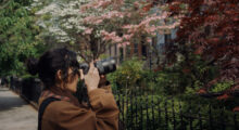 A young woman with her long dark hair tied back turns to the side and takes a picture of the front of a house, which is out of frame. She stands among trees that bloom with pink and white flowers.