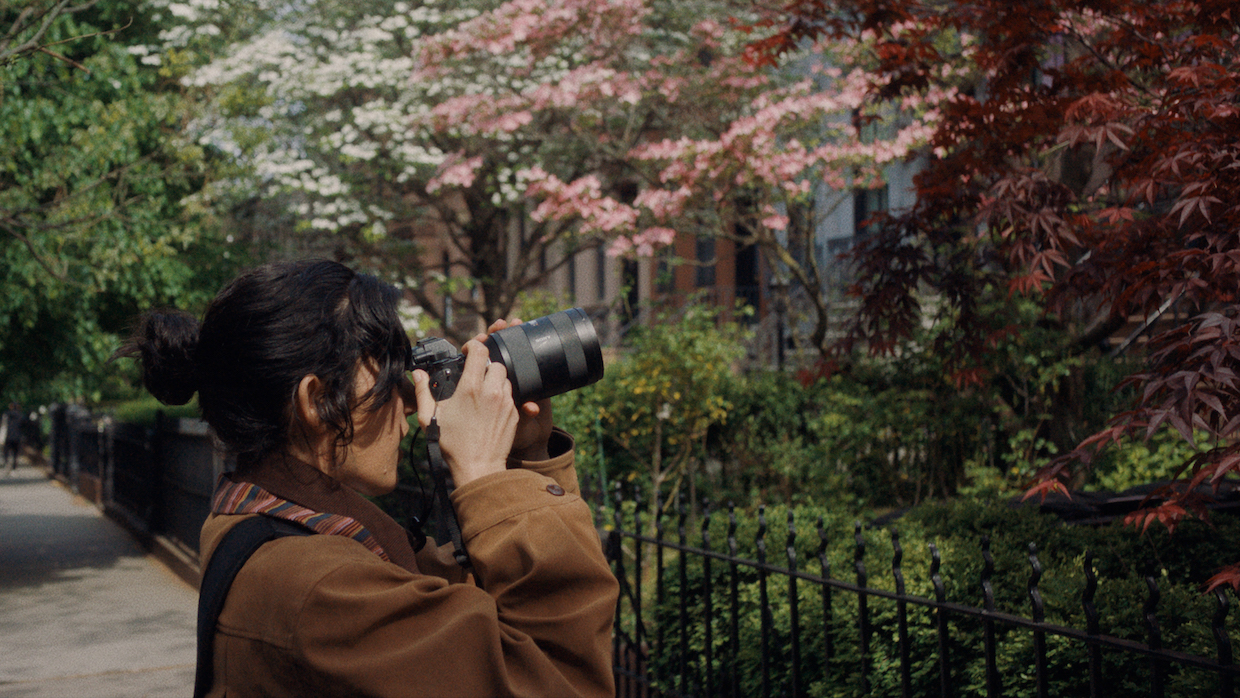 A young woman with her long dark hair tied back turns to the side and takes a picture of the front of a house, which is out of frame. She stands among trees that bloom with pink and white flowers.