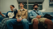 A young woman with brown hair, a tall man with salt and pepper hair and a man with a beard and afro sit together on a subway seat.