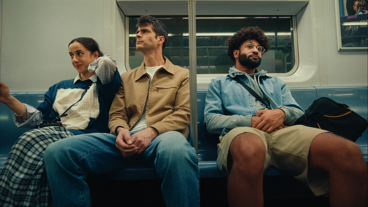 A young woman with brown hair, a tall man with salt and pepper hair and a man with a beard and afro sit together on a subway seat.