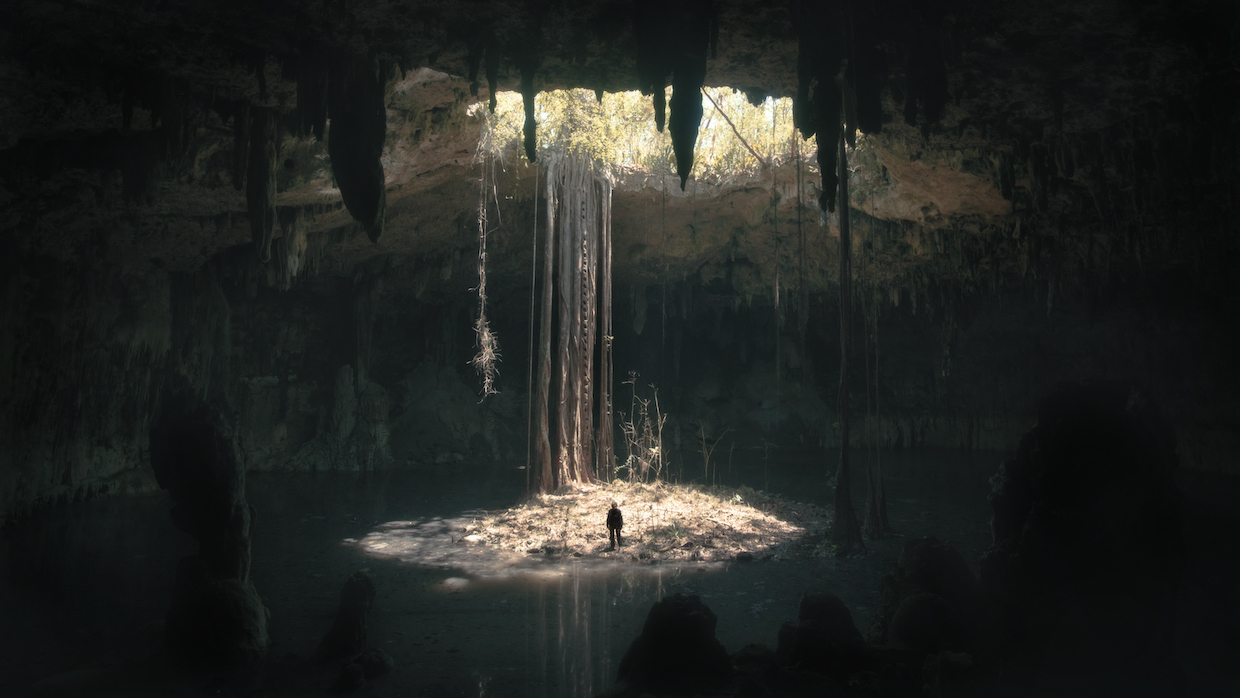 A beam of sunlight radiates town into a dark cavern, where a person stands and looks incredibly small amid the massive, dark space.