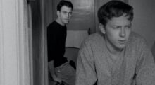 A black and white image of one man looking out of a window while another man leans on the wall behind him and looks in the same direction.