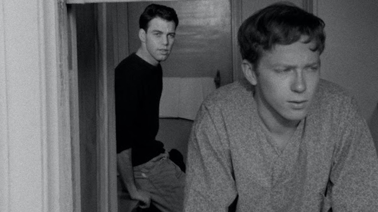A black and white image of one man looking out of a window while another man leans on the wall behind him and looks in the same direction.