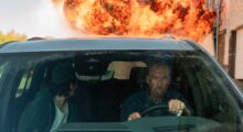 An angry man covered in blood drives away from a fireball.