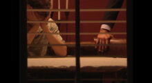 A man holds his head in his hands while a man rests his hands on a chair next to him.