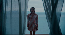 A young woman wearing a red and white dress with red and white polkadots stands between sheer grey curtains, her back to the vast blue sea behind her.