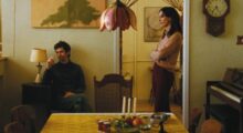 A man sits smoking a cigarette on a chair while a woman stands next to him.