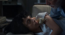 A young woman with curly brown hair lays on her side as an older man with white hair holds a phone to her ear.