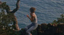 A woman wearing blue jeans, a white tank top and her hair in a bun smokes a cigarette and overlooks the ocean.